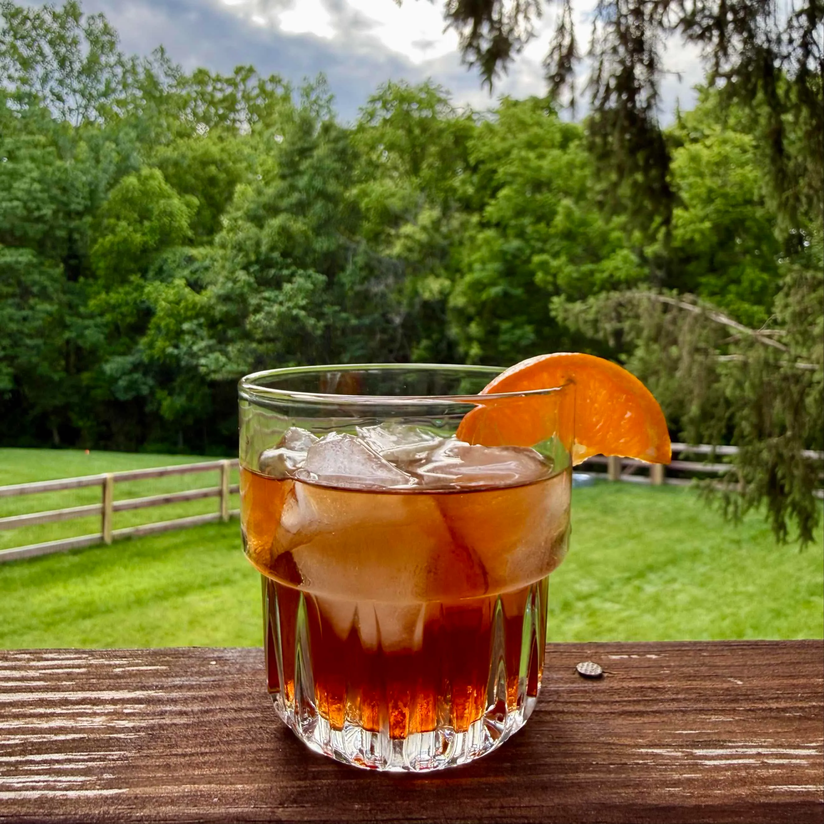 Berry Barron cocktail, a deep amber bourbon and Chambord old fashioned over ice in a rocks glass with an orange slice, on a deck rail overlooking a green backyard
